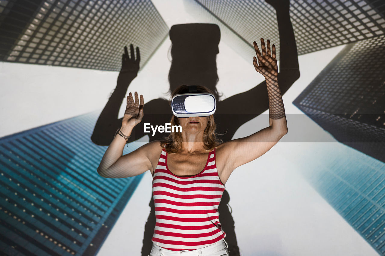 Unrecognizable female in casual wear and vr headset standing with arms raised against contemporary skyscrapers inside virtual city
