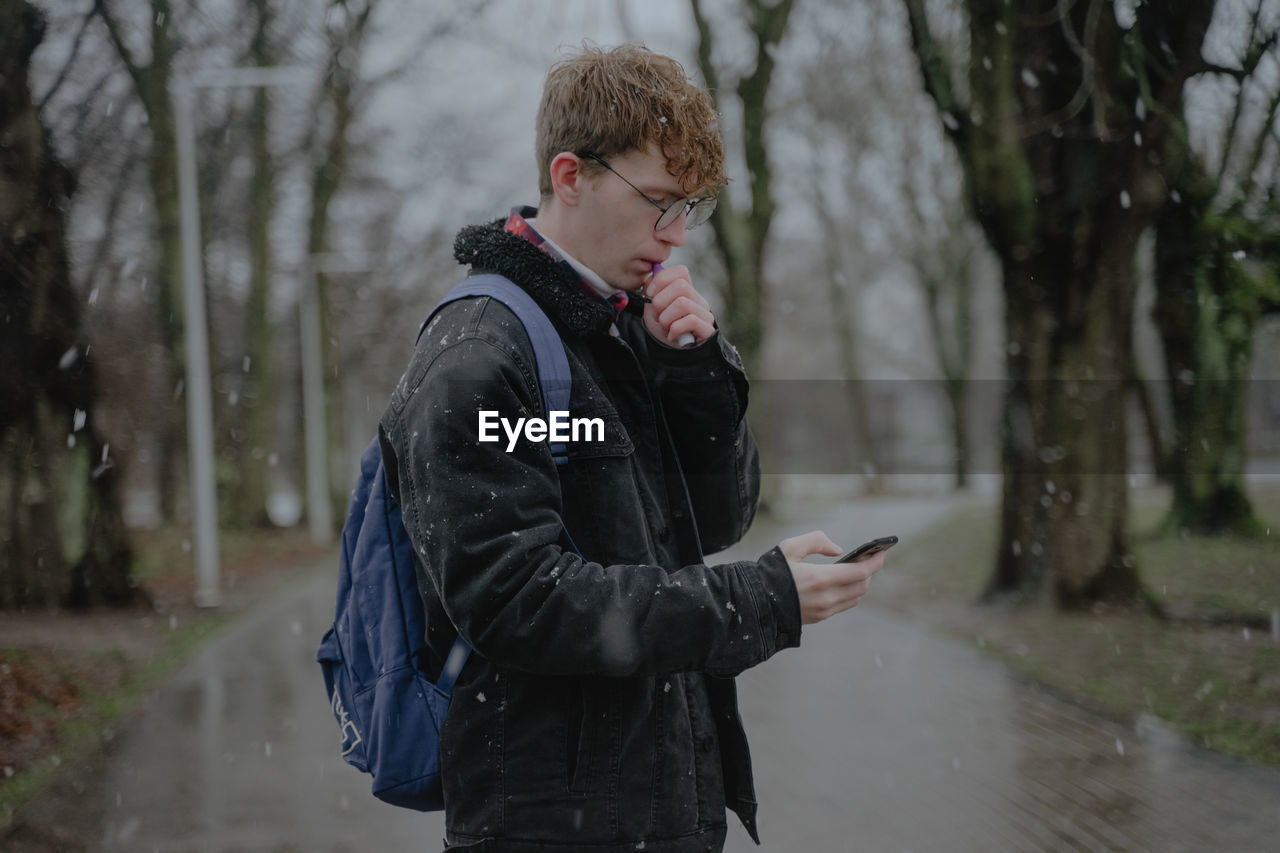 It's snowing, a guy in the park smokes an electronic cigarette and looks at a mobile phone