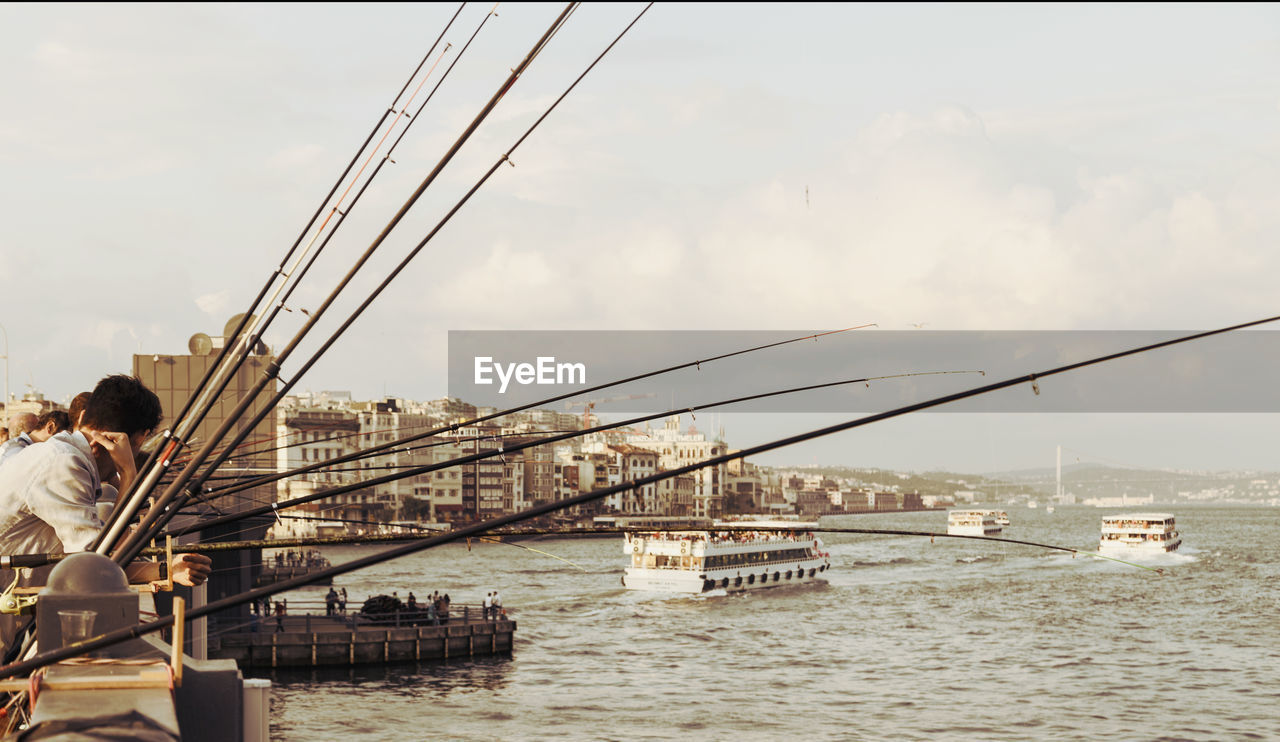 People with fishing rod by sea against sky in city