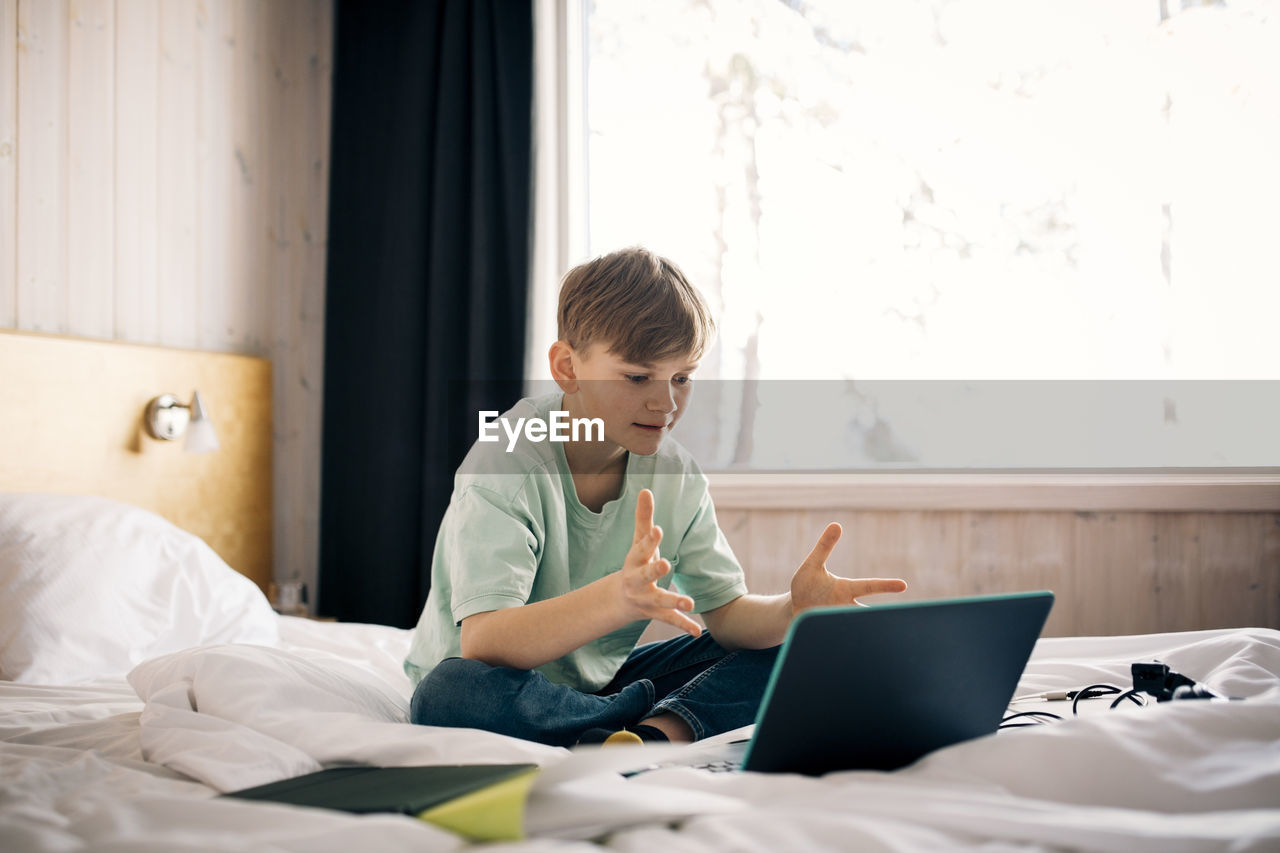 Boy attending video call while gesturing during homeschooling in bedroom