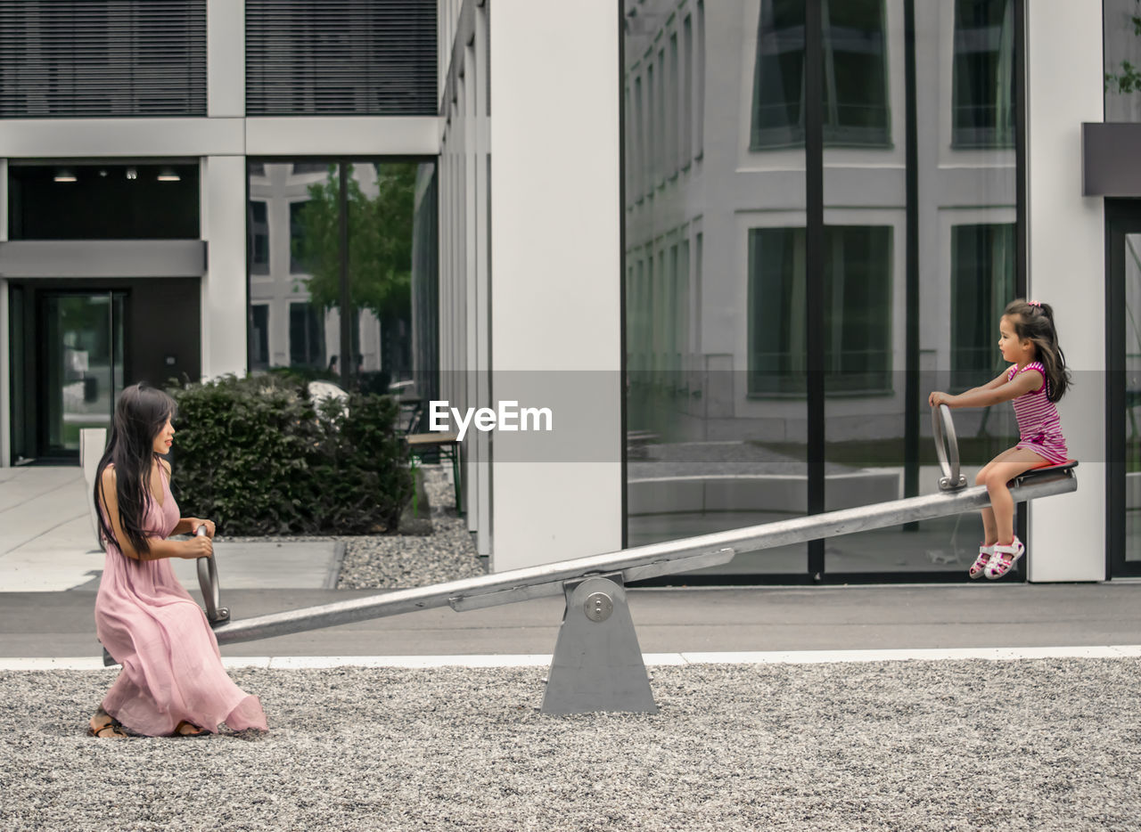 Side view of mother with daughter playing on seesaw against building