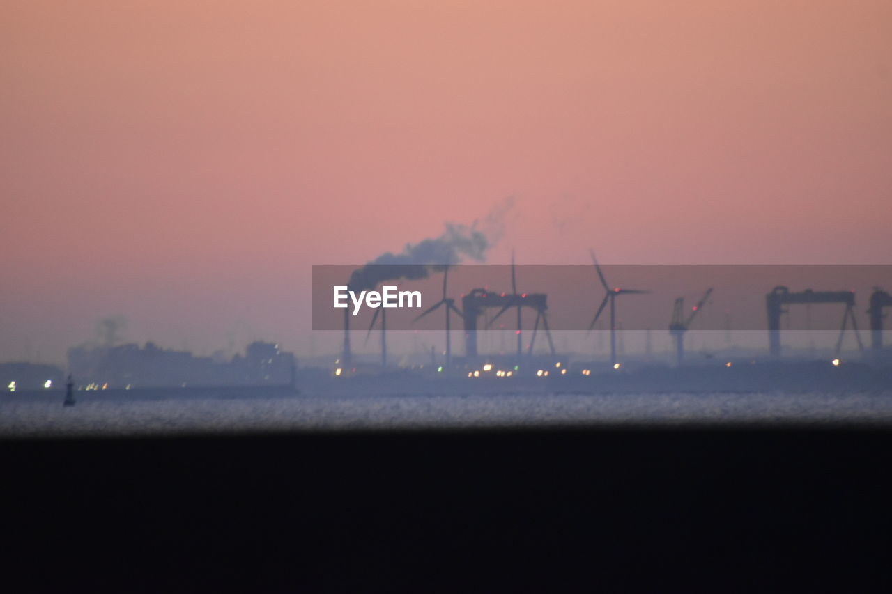 SILHOUETTE OF FACTORY BY SEA AGAINST ORANGE SKY