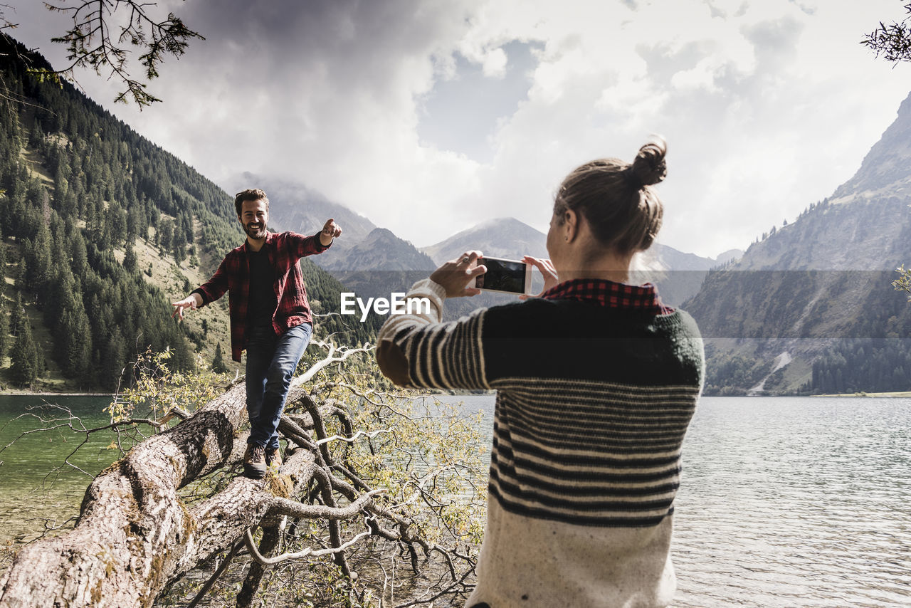 Austria, tyrol, alps, woman taking cell phone picture of man balancing on tree trunk at mountain lake