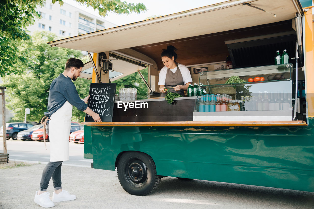 Male owner adjusting board on concession stand while partner working in food truck