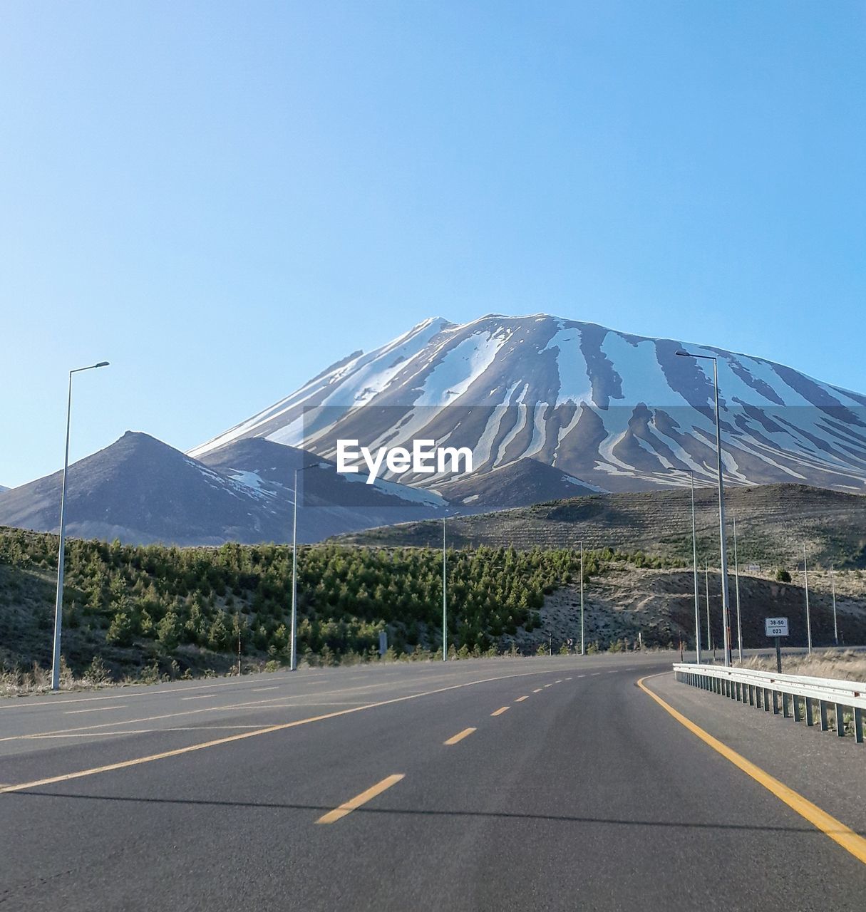 road, mountain, sky, transportation, highway, nature, no people, clear sky, day, road marking, environment, scenics - nature, beauty in nature, marking, landscape, blue, symbol, travel, travel destinations, sunny, freeway, city, outdoors, land, sign, tranquility, architecture, the way forward, infrastructure, tranquil scene, copy space, street, asphalt, built structure, empty, road trip, mountain range, absence, sunlight, empty road, diminishing perspective