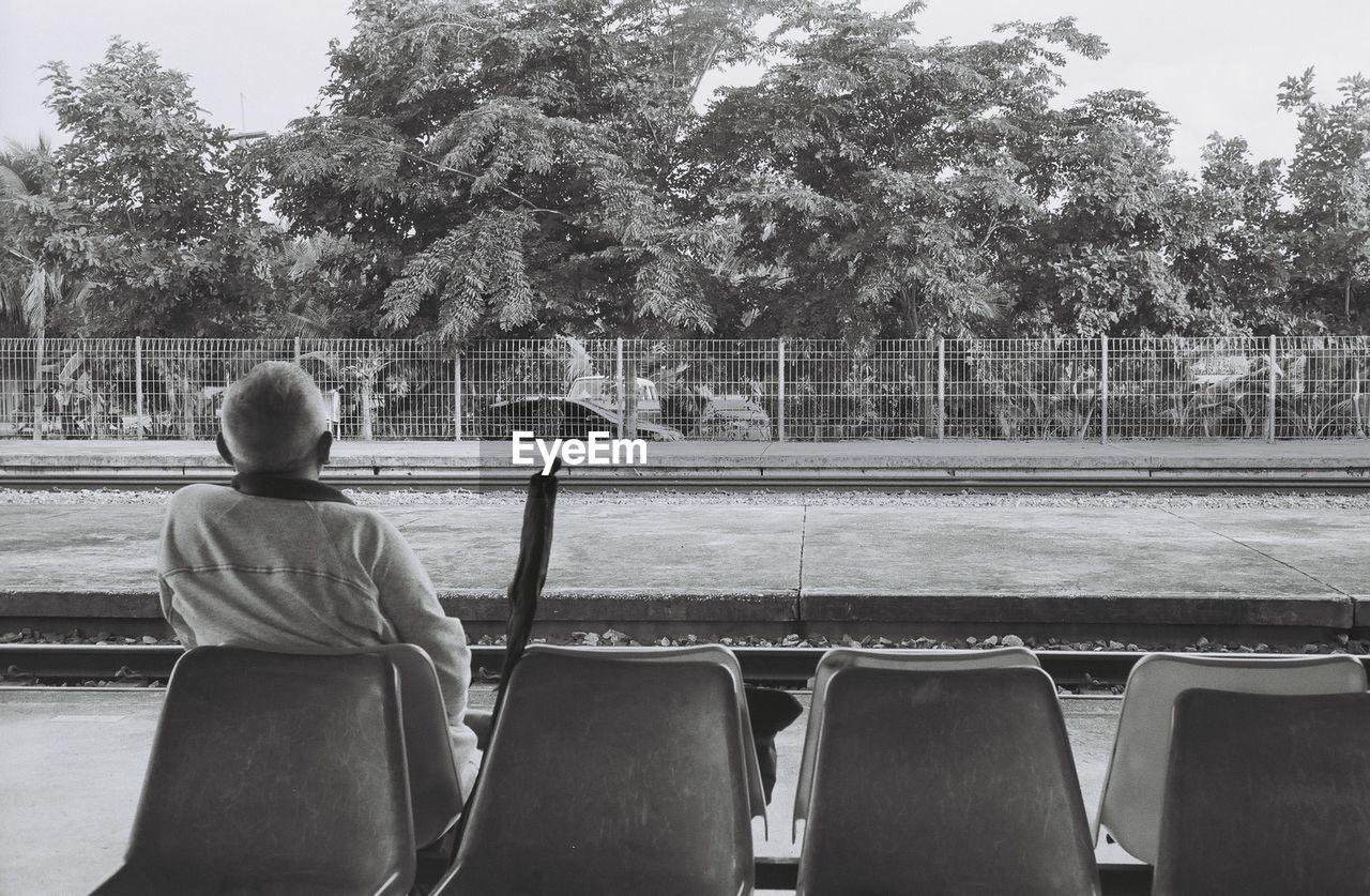 Rear view of senior man sitting at railroad station platform against trees