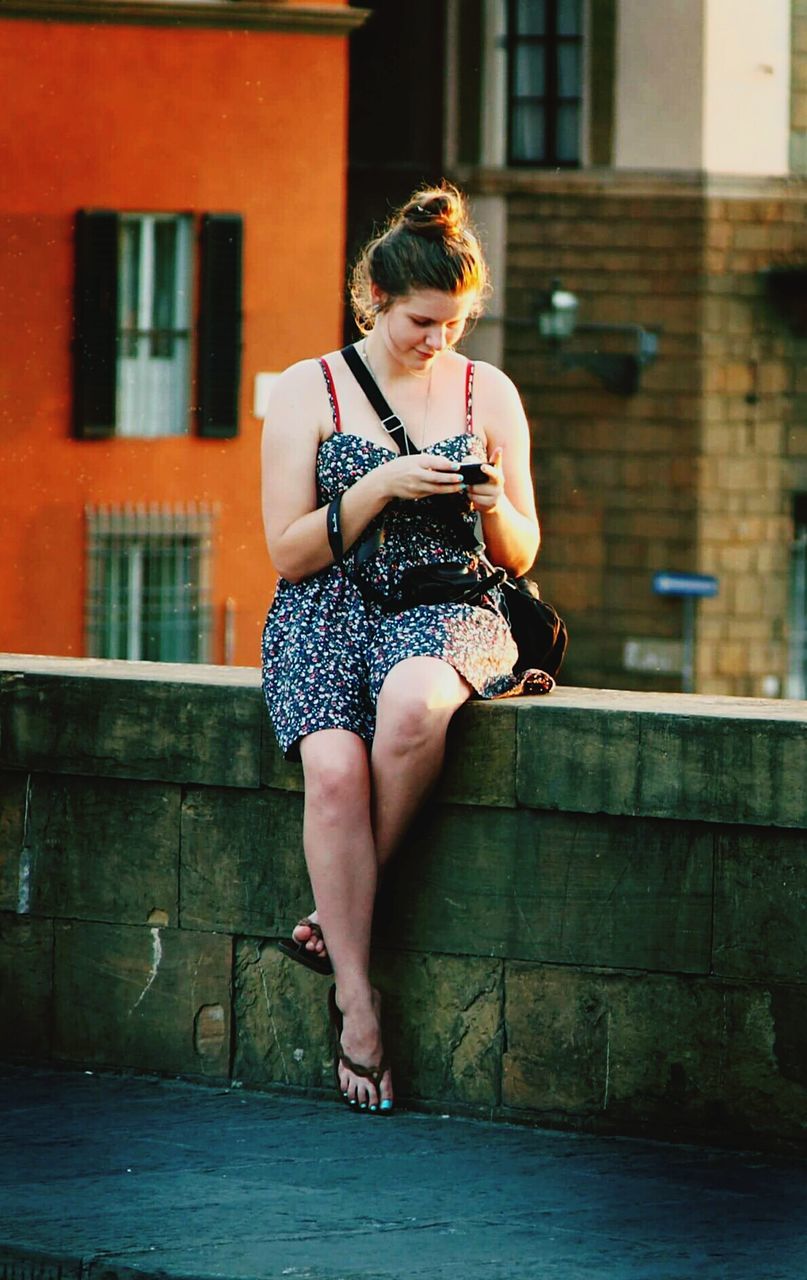 Full length of woman using phone while sitting on retaining wall against building