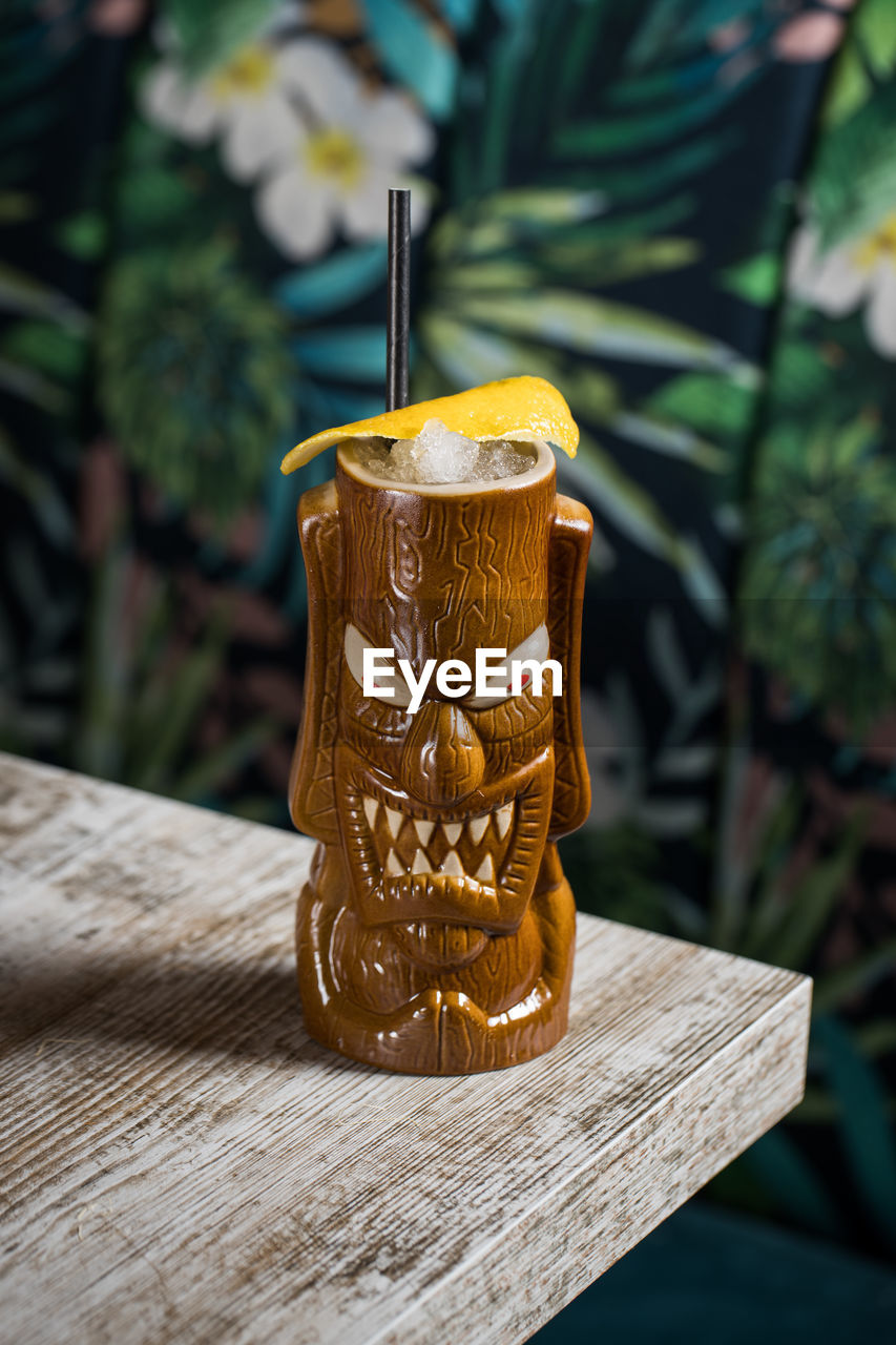 Brown sculptural tiki mug with alcohol drink decorated with straw and ice placed on table on blurred background