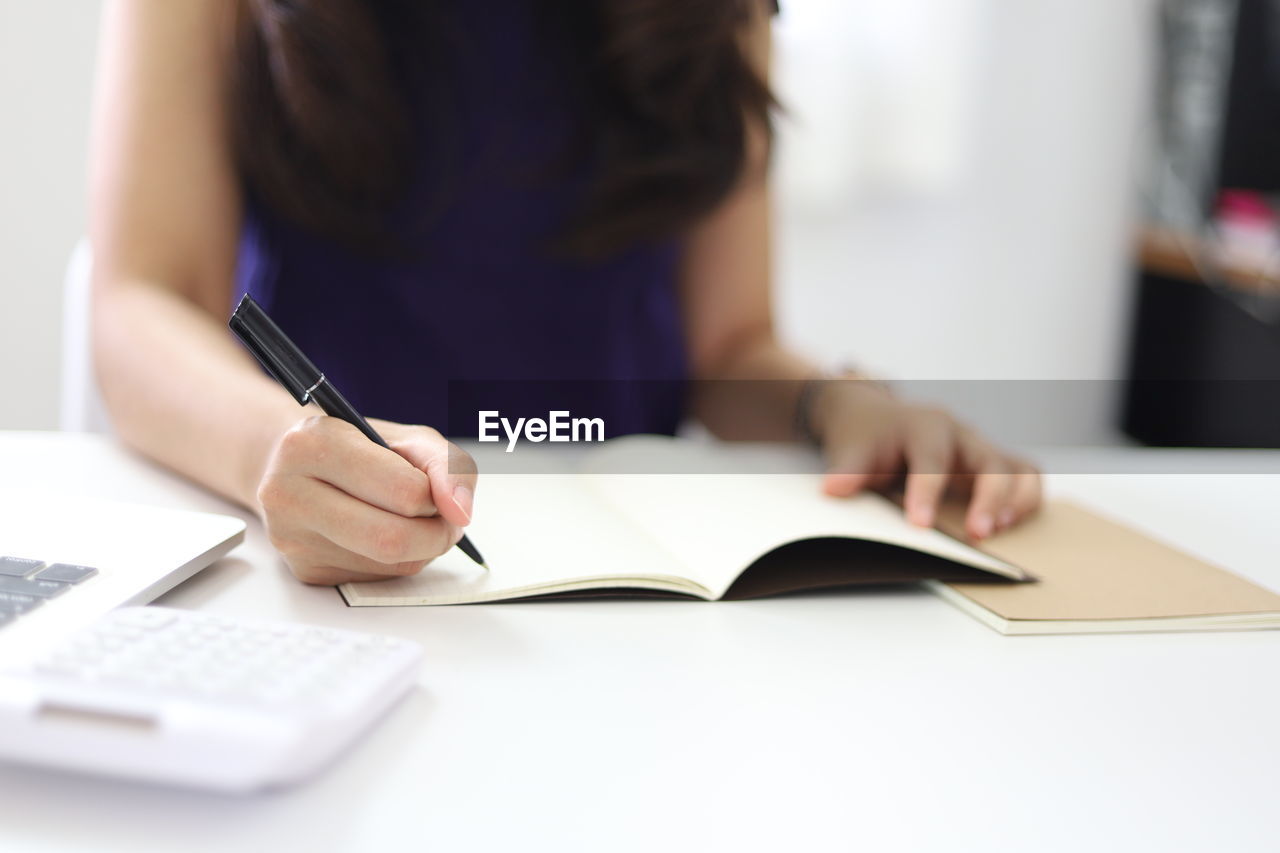 MIDSECTION OF WOMAN READING BOOK WHILE SITTING ON TABLE AT HOME