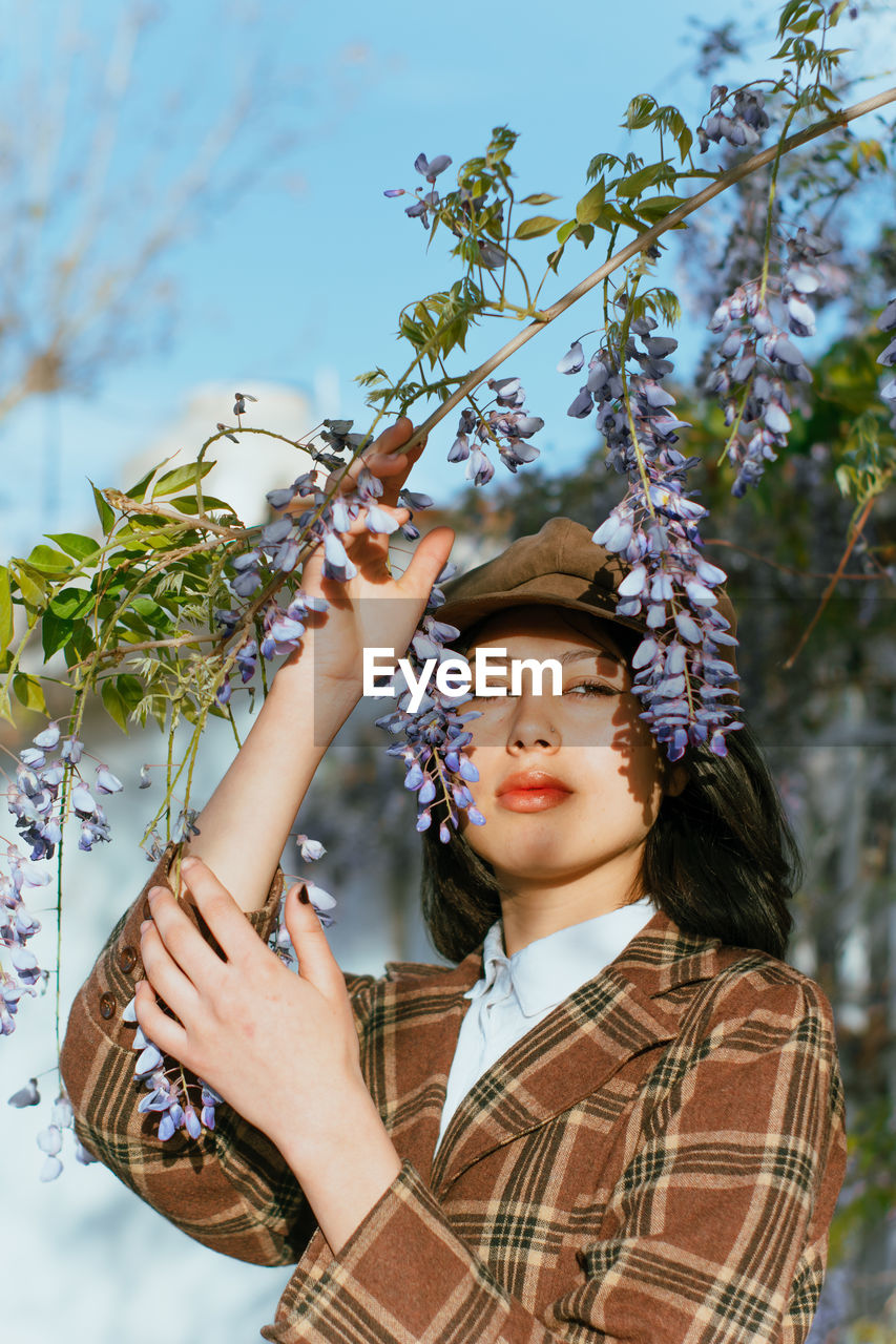 Young gentle ethnic female in checkered wear with beret touching blossoming plant while looking away on street