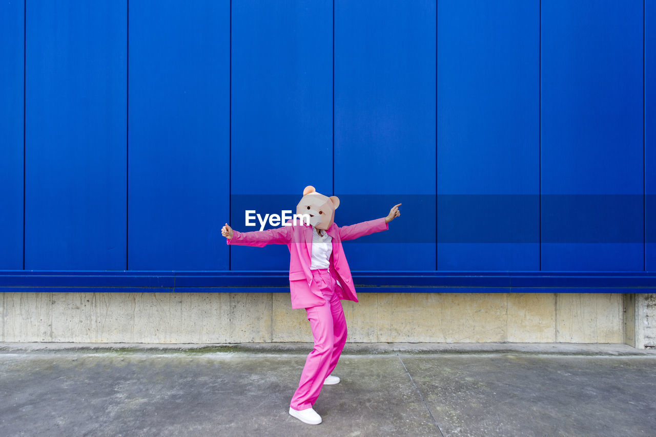 Woman wearing vibrant pink suit and bear mask standing with outstretched arms in front of blue wall