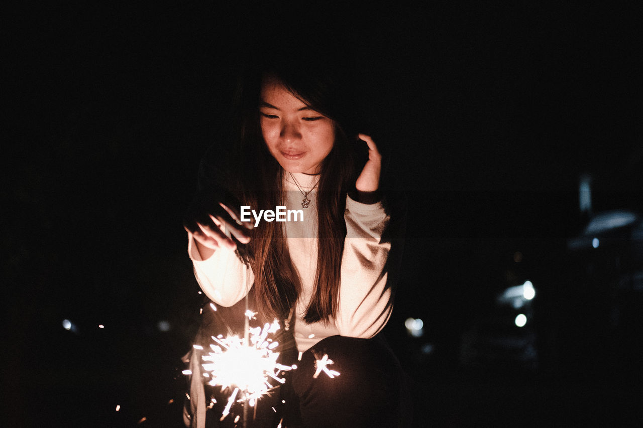 Young woman holding illuminated sparkler while sitting at night