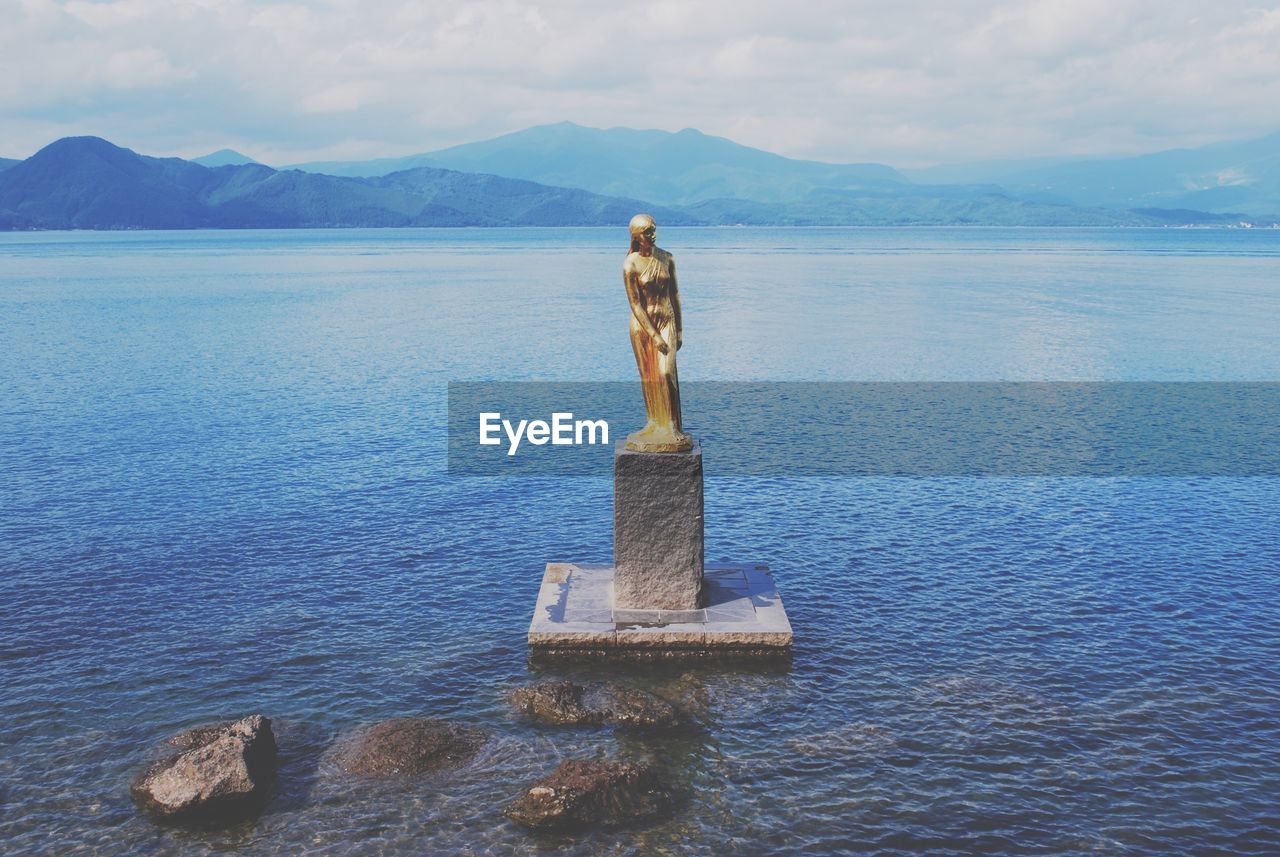 Gold female statue in lake against sky