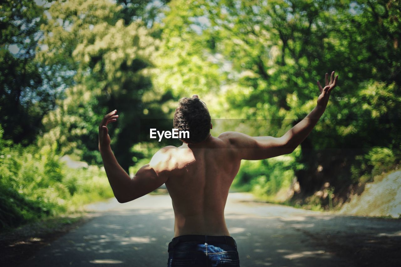 Rear view of shirtless man gesturing while standing on road amidst trees
