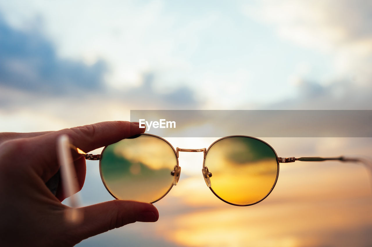 Hand holds metal round sunglasses through which you can see sea sunset in blue and orange colors