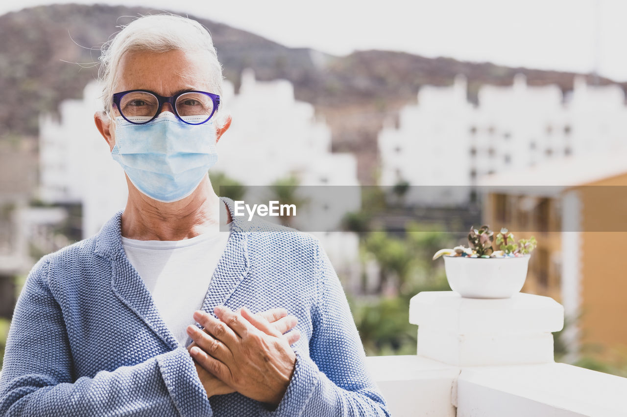 Portrait of senior woman wearing mask with hands on chest standing against building