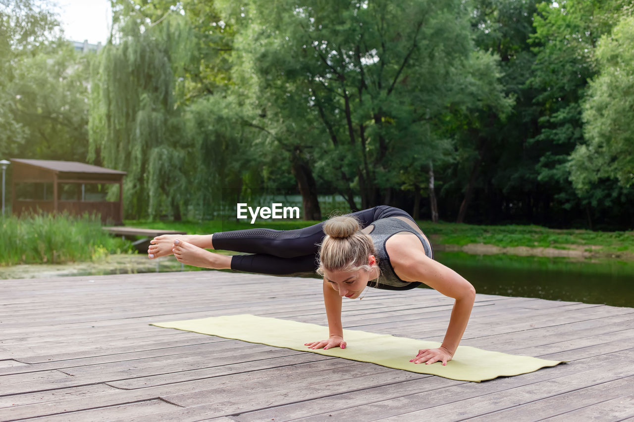 A beautiful woman on a wooden platform by a pond on mat in summer, does yoga