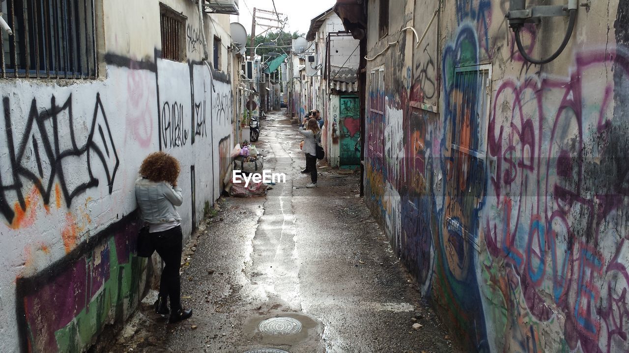 People photographing in wet alley amidst graffiti walls