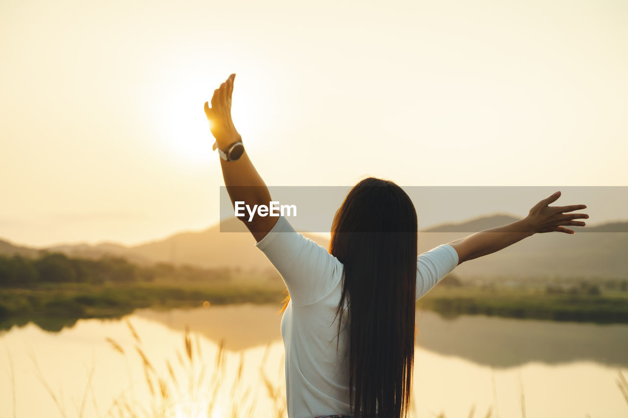 Rear view of woman with arms raised standing against sky during sunset