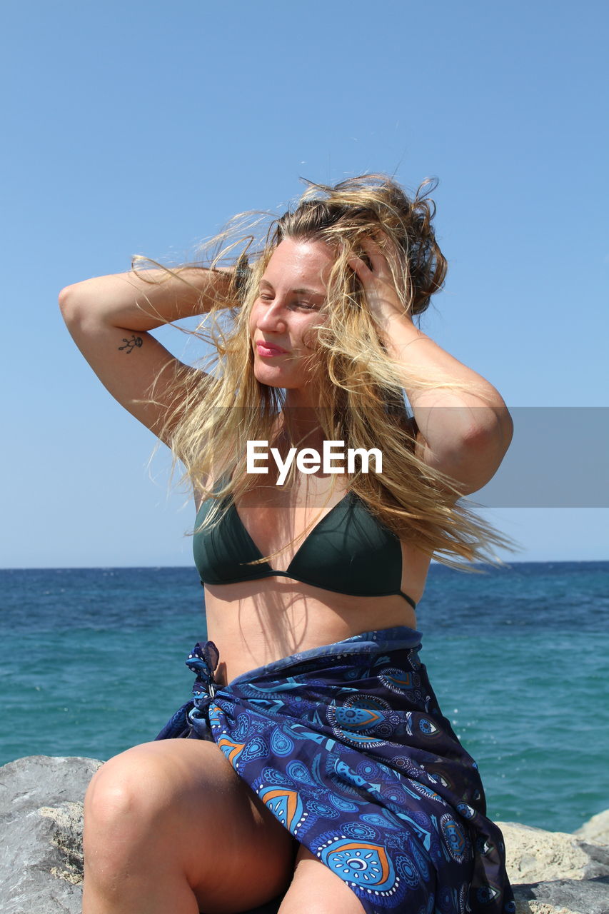 Young woman sitting on rock at beach against clear sky