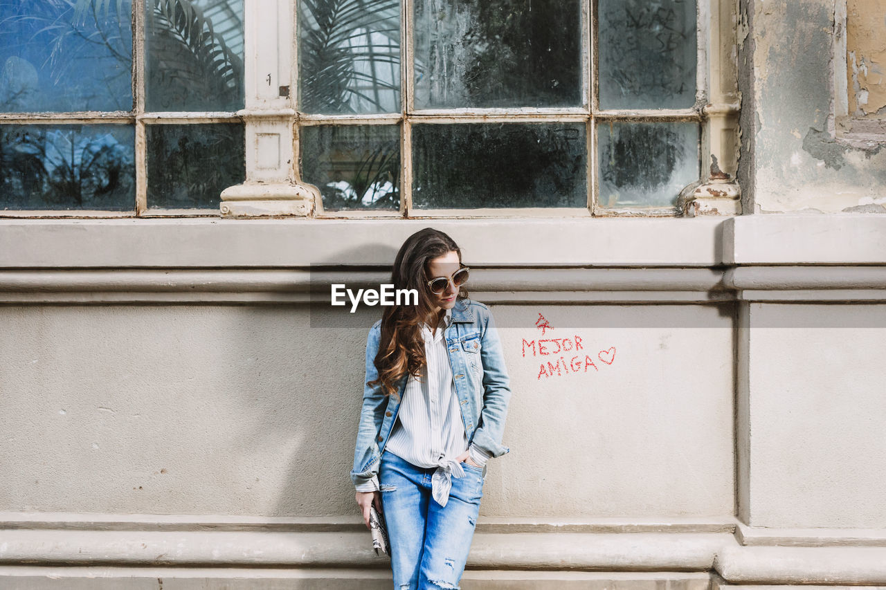 Young female in trendy denim outfit and sunglasses standing with hand in pocket near shabby building in city