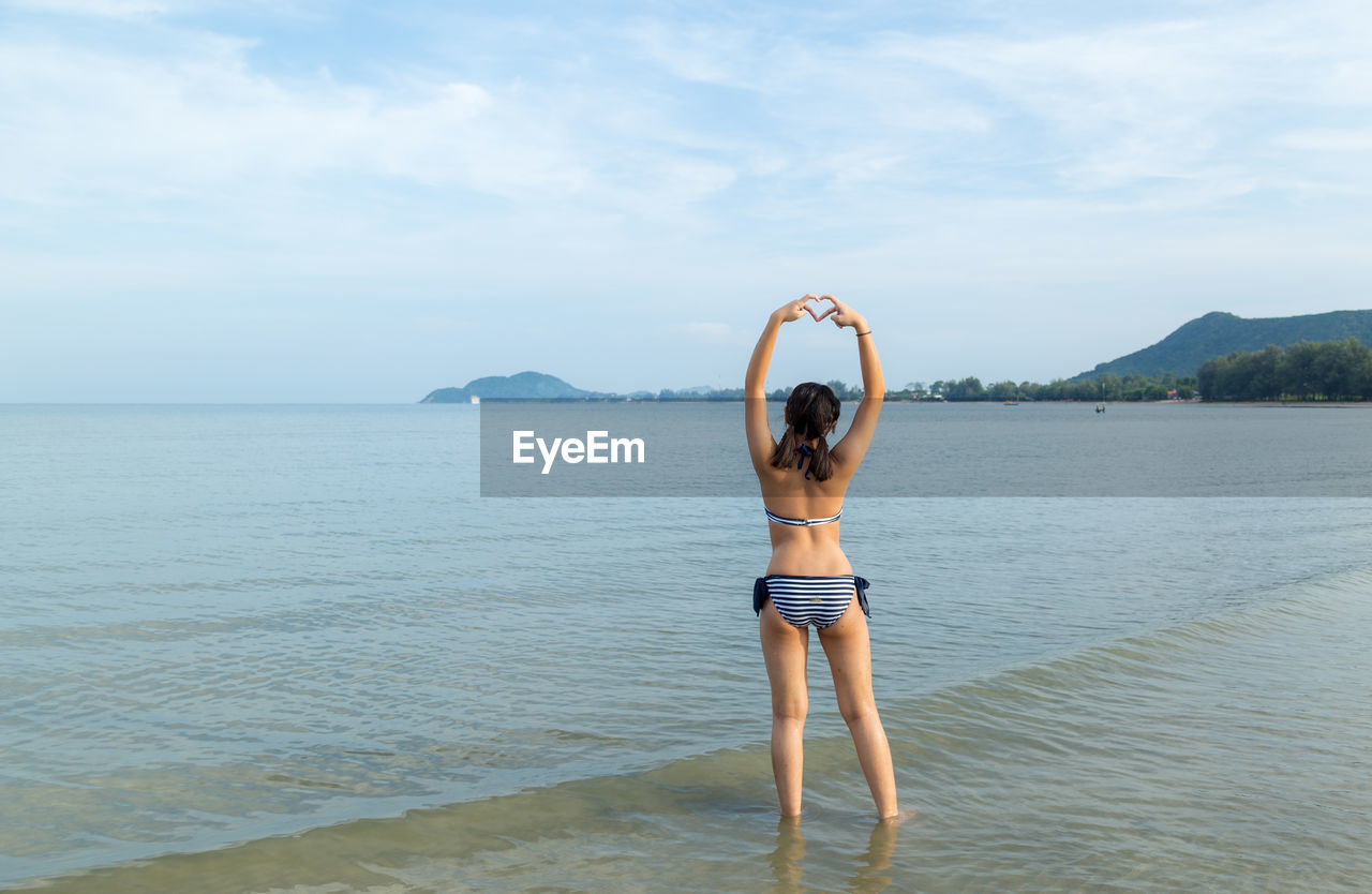 Girl making heart shape at beach against sky