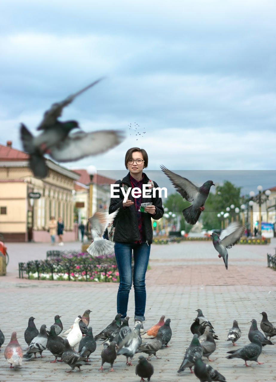 Woman teenager feeding pigeons with seeds on wide pedestrian city street, urban bird catches food