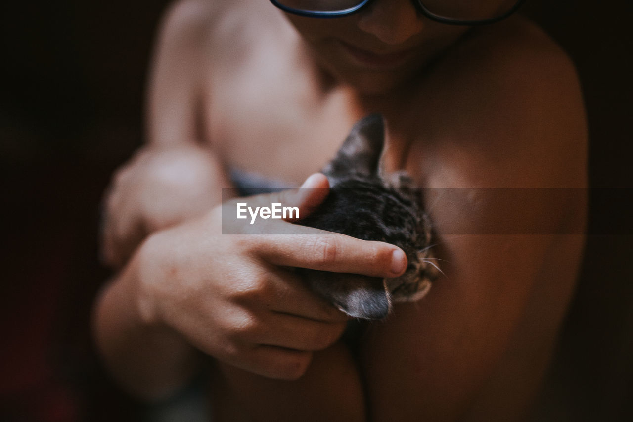 Midsection of shirtless teenage boy holding cat against black background