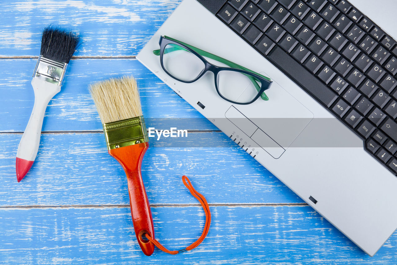 High angle view of laptop with eyeglasses and paintbrushes on blue wooden table