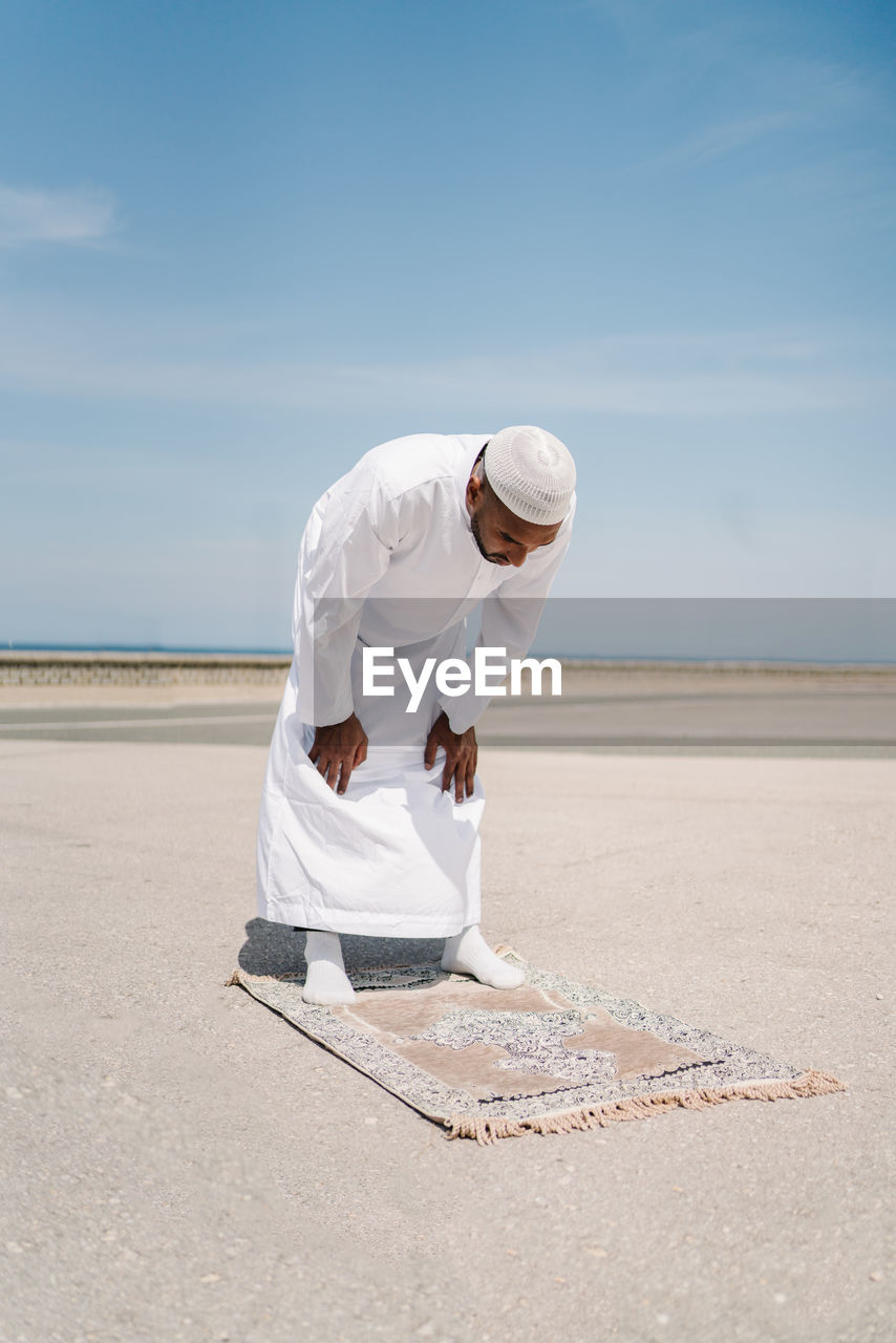 Full boy islamic male in traditional white clothes standing on rug and praying against blue sky on beach