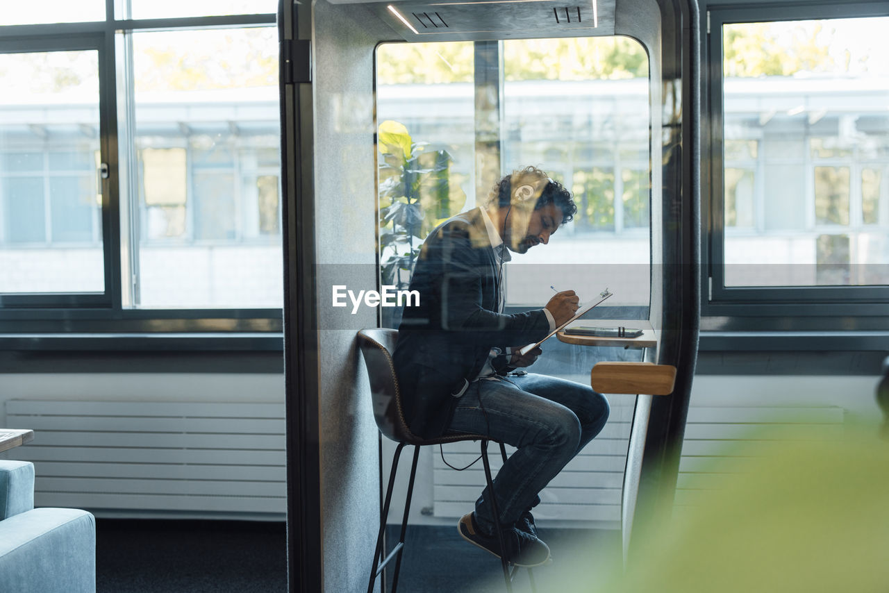 Businessman writing on clipboard in soundproof cabin at office