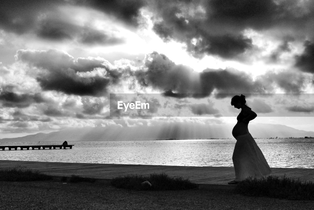 Pregnant woman standing on shore at beach against cloudy sky