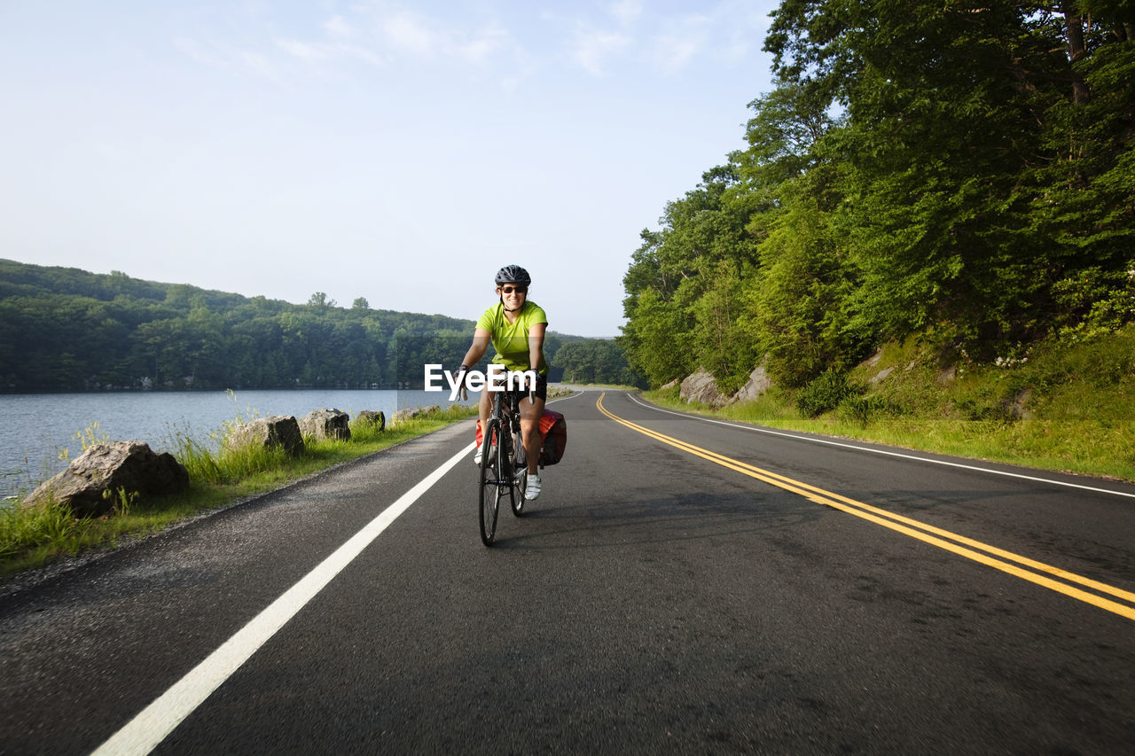 Woman cycling on road amidst trees and lake against sky