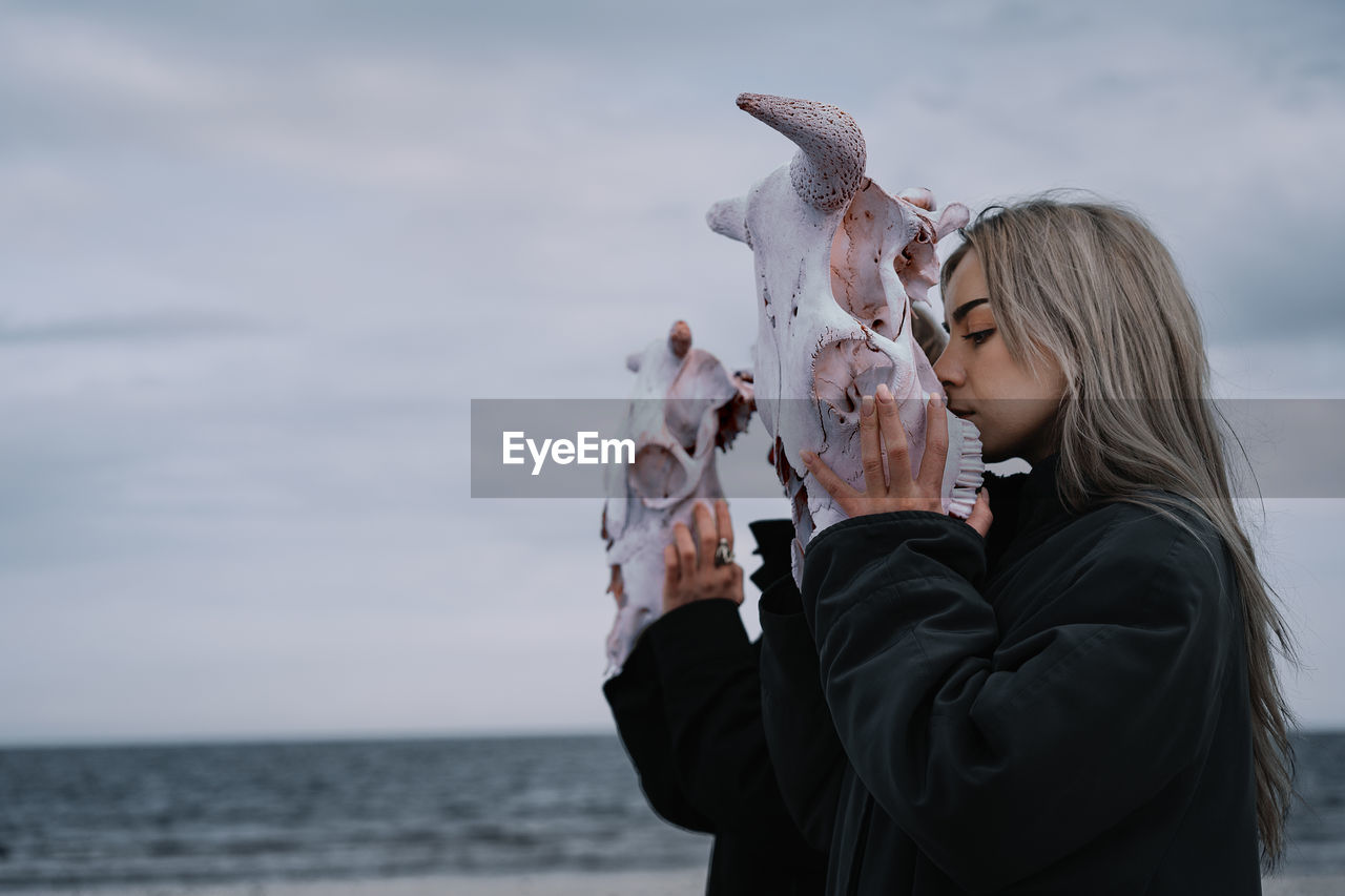 Woman holding cow skull by the sea against cloudy sky