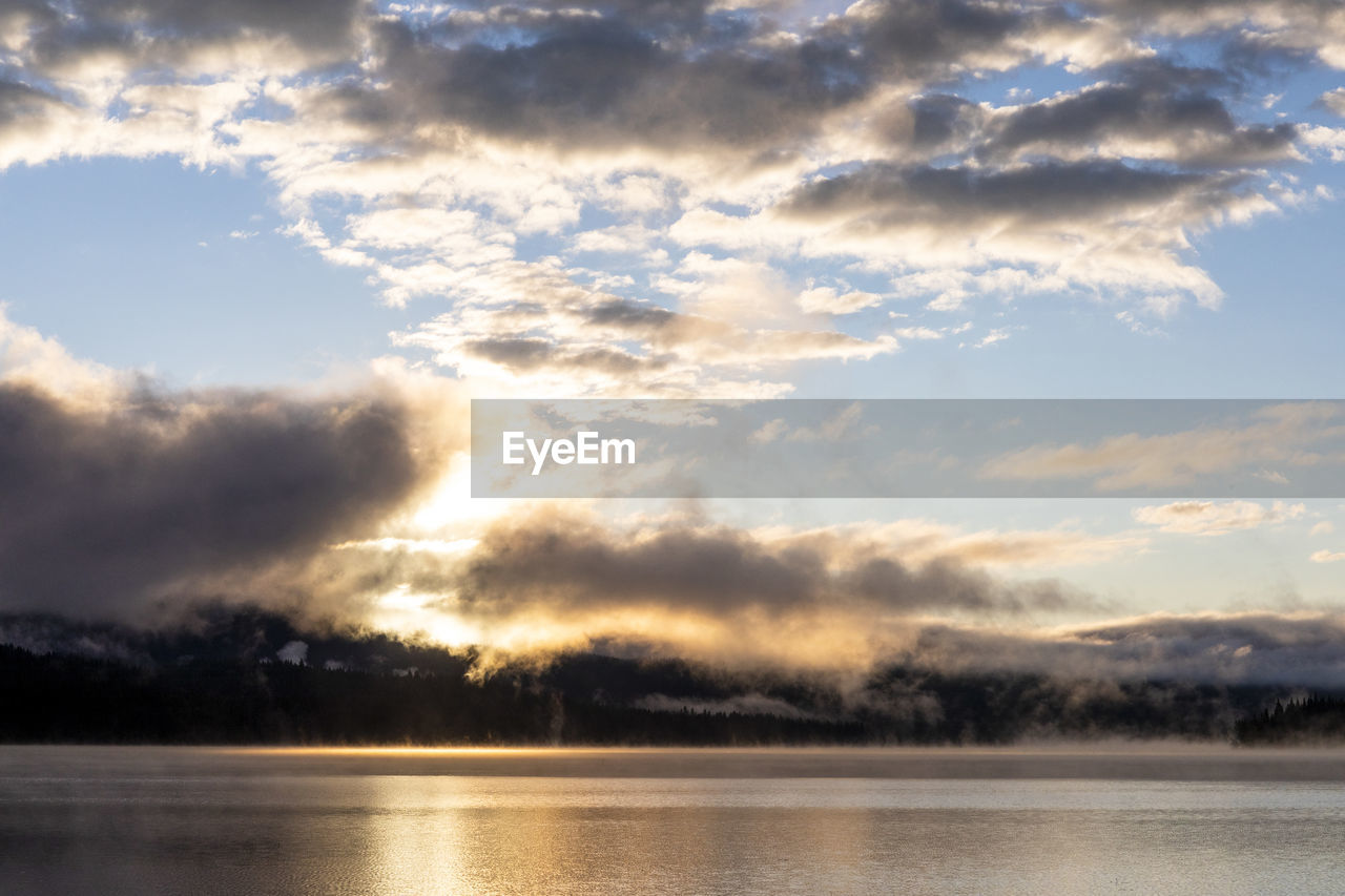Sunrise over calm lake water with clouds and trees in countryside
