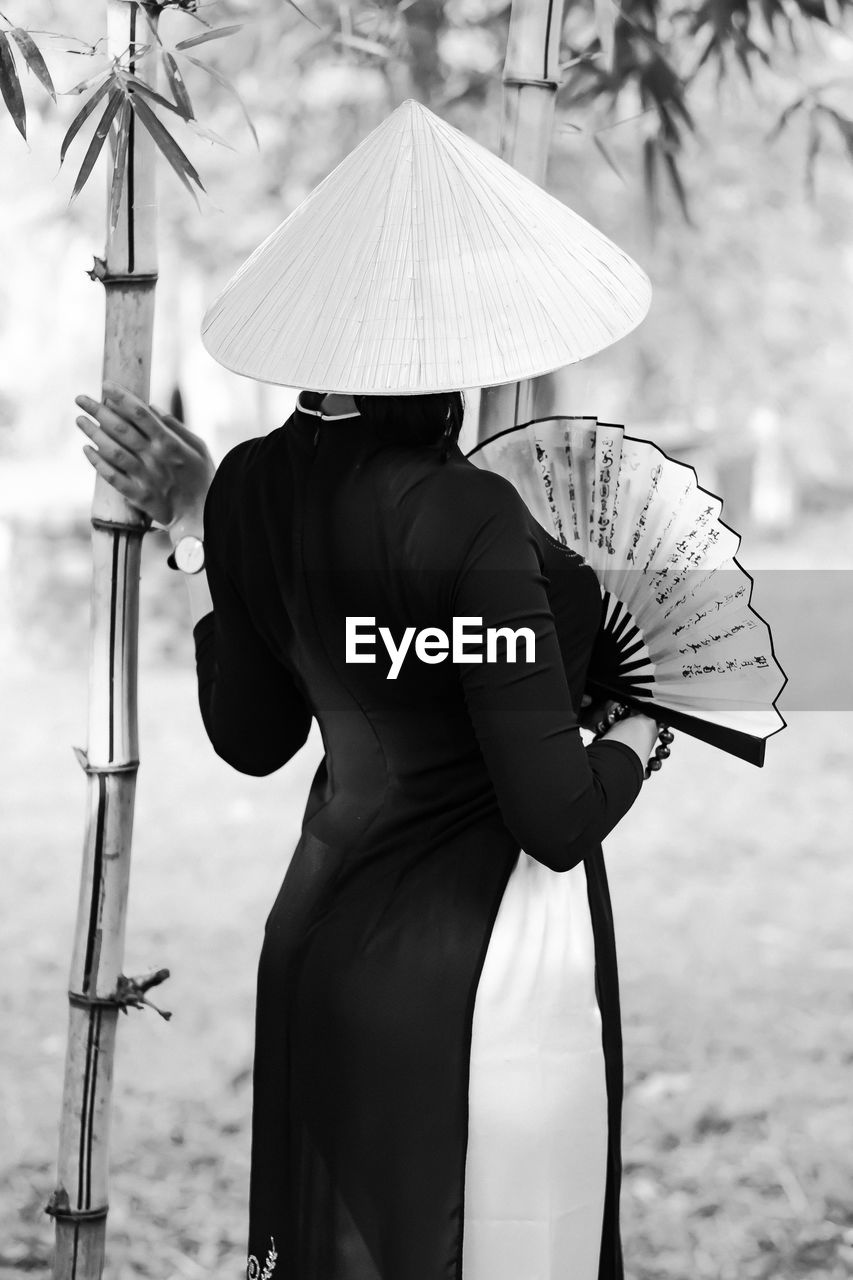 Rear view of woman holding hand fan while standing by bamboo plant