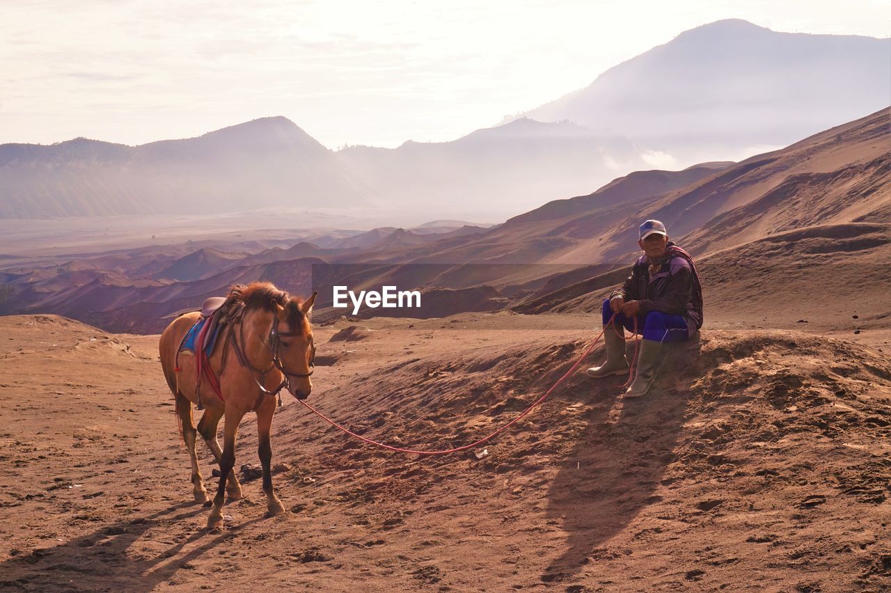 Full length of man sitting by horse at desert against mountains