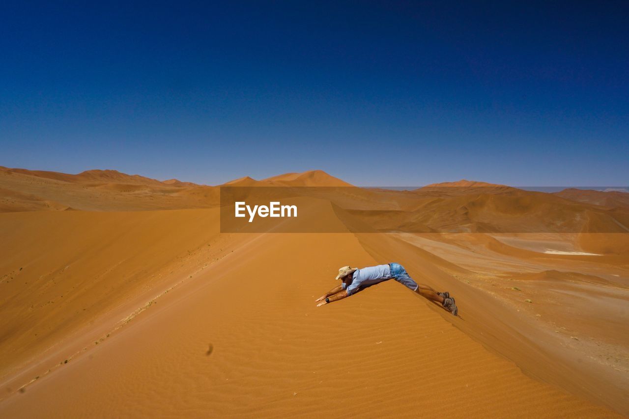 Side view of man lying on sand dune in desert against clear blue sky