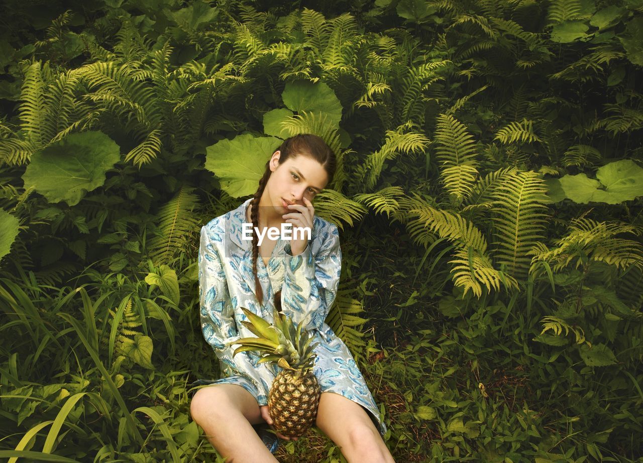 Portrait of young woman holding pineapple while sitting against plants