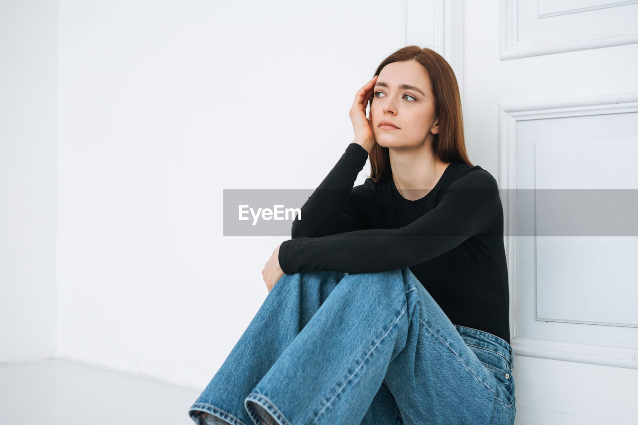 Young unhappy woman girl with long hair in jeans on floor in closed position by door at home