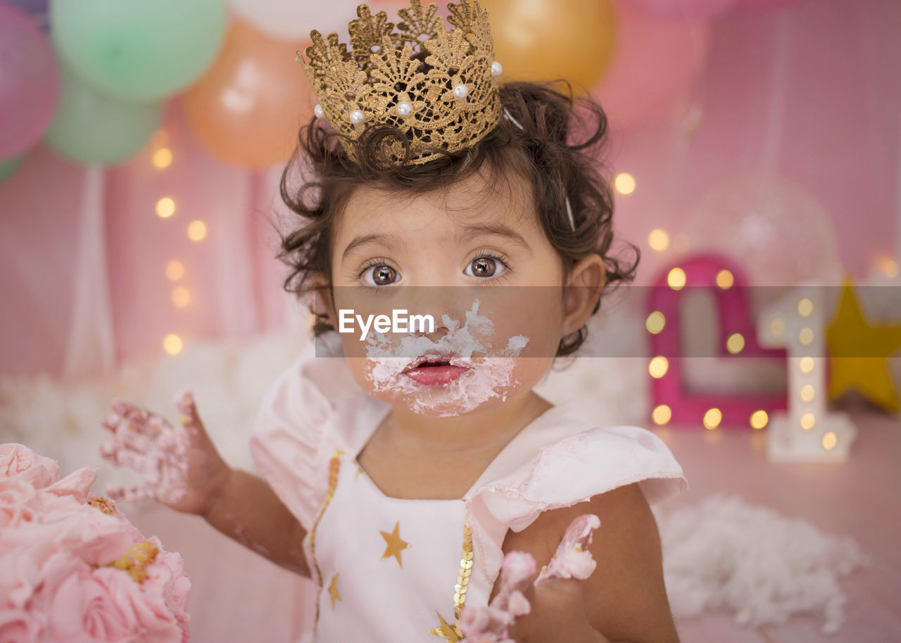 Cute baby girl with balloons and birthday cake sitting at home