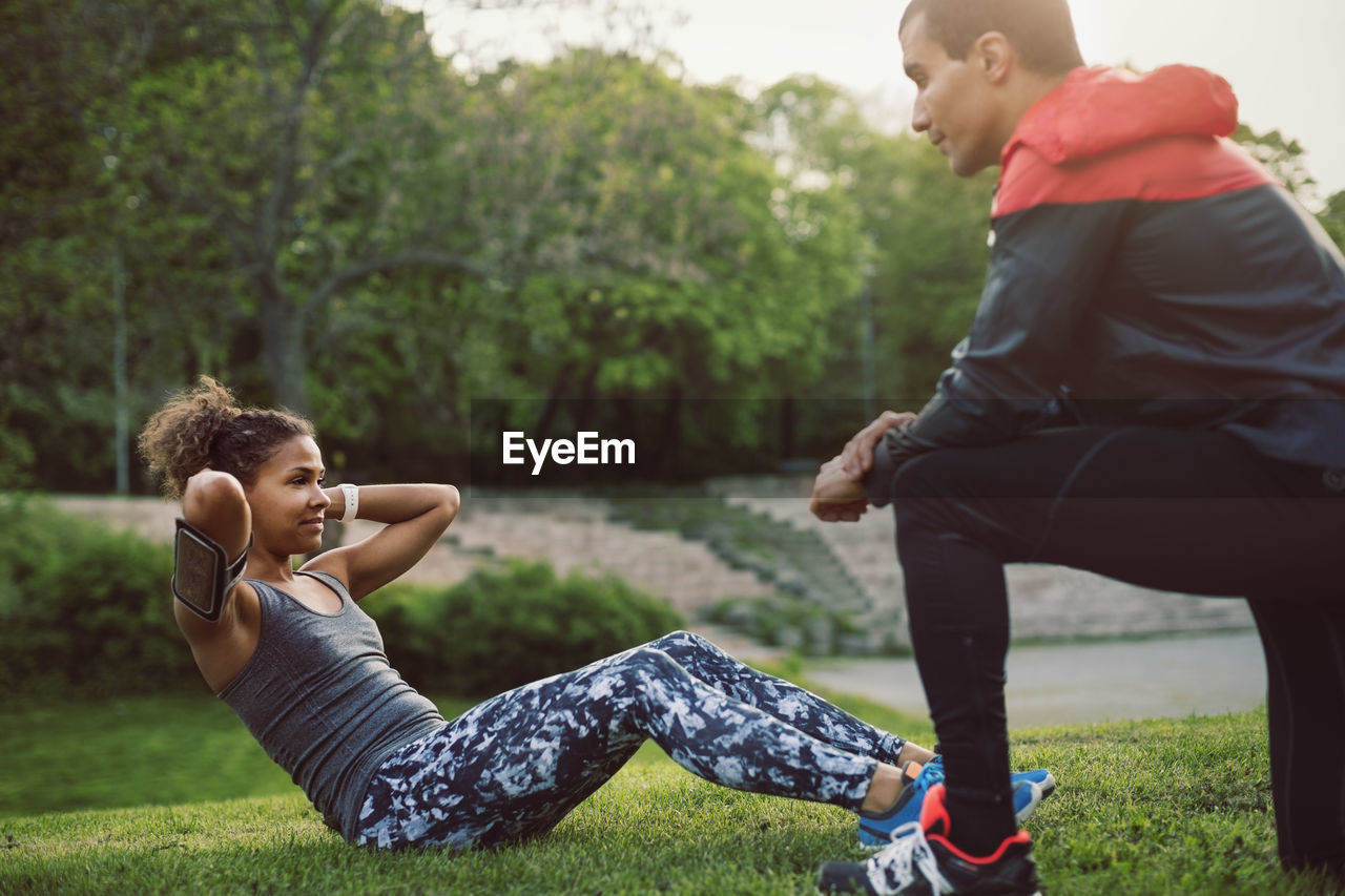 Man kneeling and looking at woman doing sit-ups on field