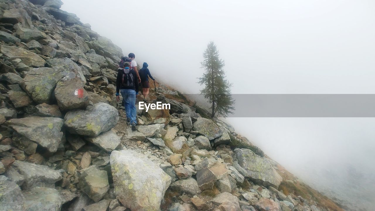 REAR VIEW OF HIKER STANDING ON ROCKS AGAINST SKY DURING FOGGY WEATHER