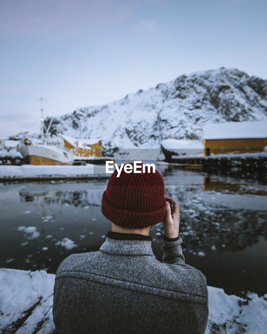 Rear view of man photographing lake while standing against sky during winter