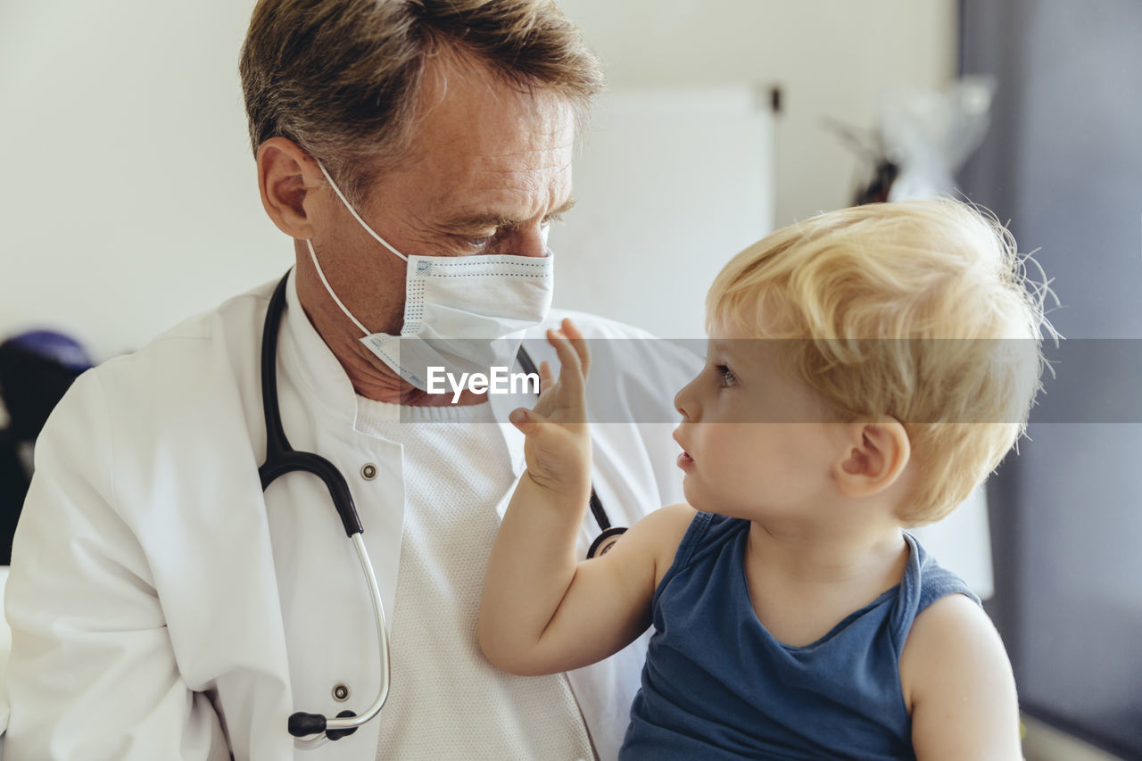 Toddler sitting on lap of pediatrician, wearing protective mask