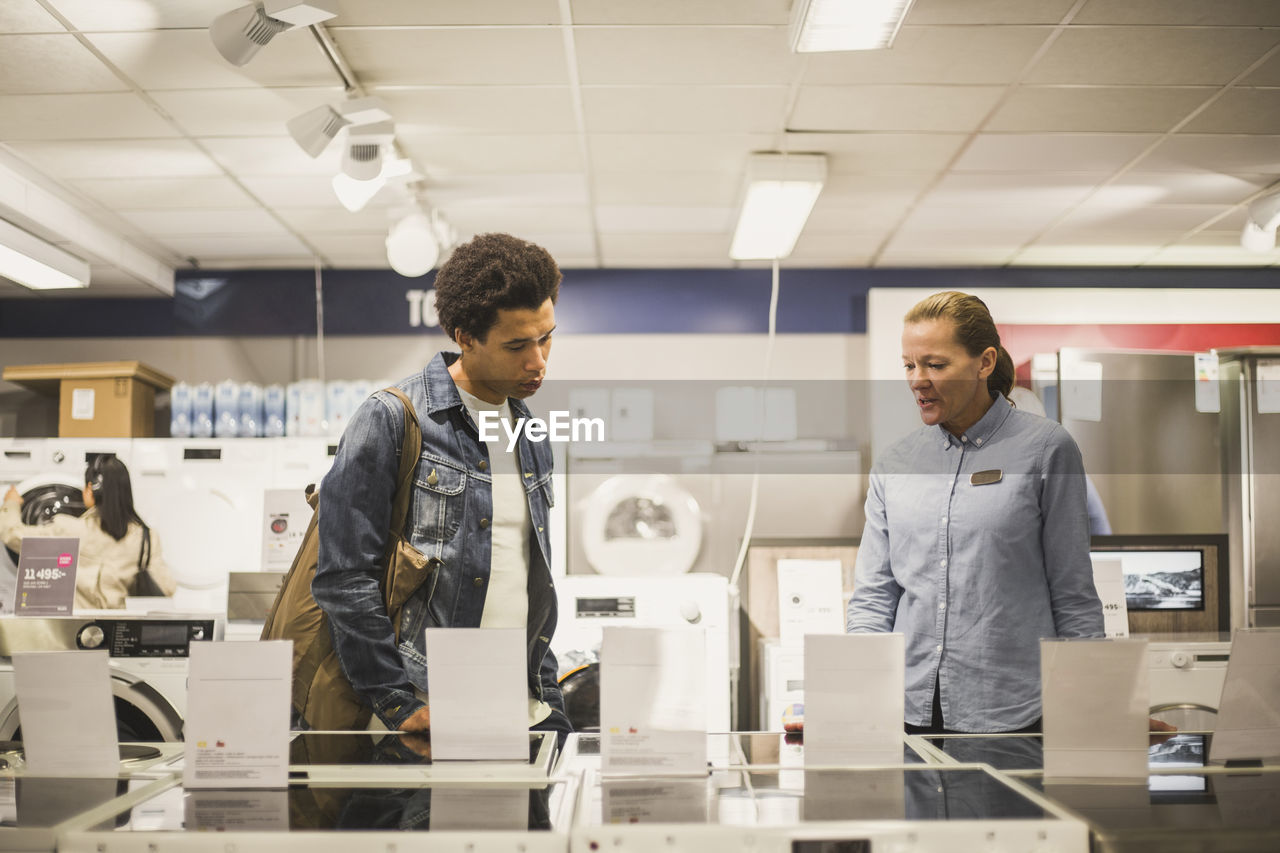 Mature female owner explaining about appliance to young customer while standing in electronics store