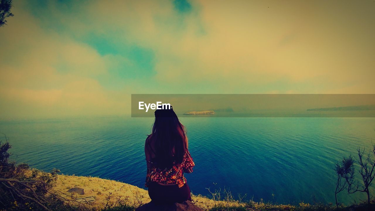 Rear view of woman sitting by sea against sky