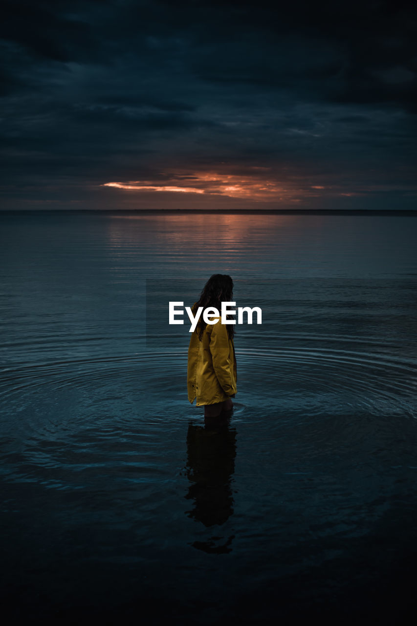 Woman standing on sea against sky during sunset