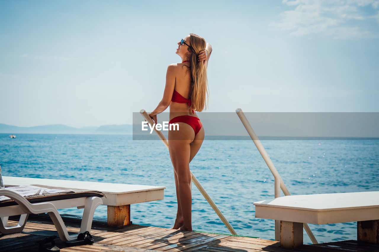 Young woman wearing red bikini while standing by sea against sky