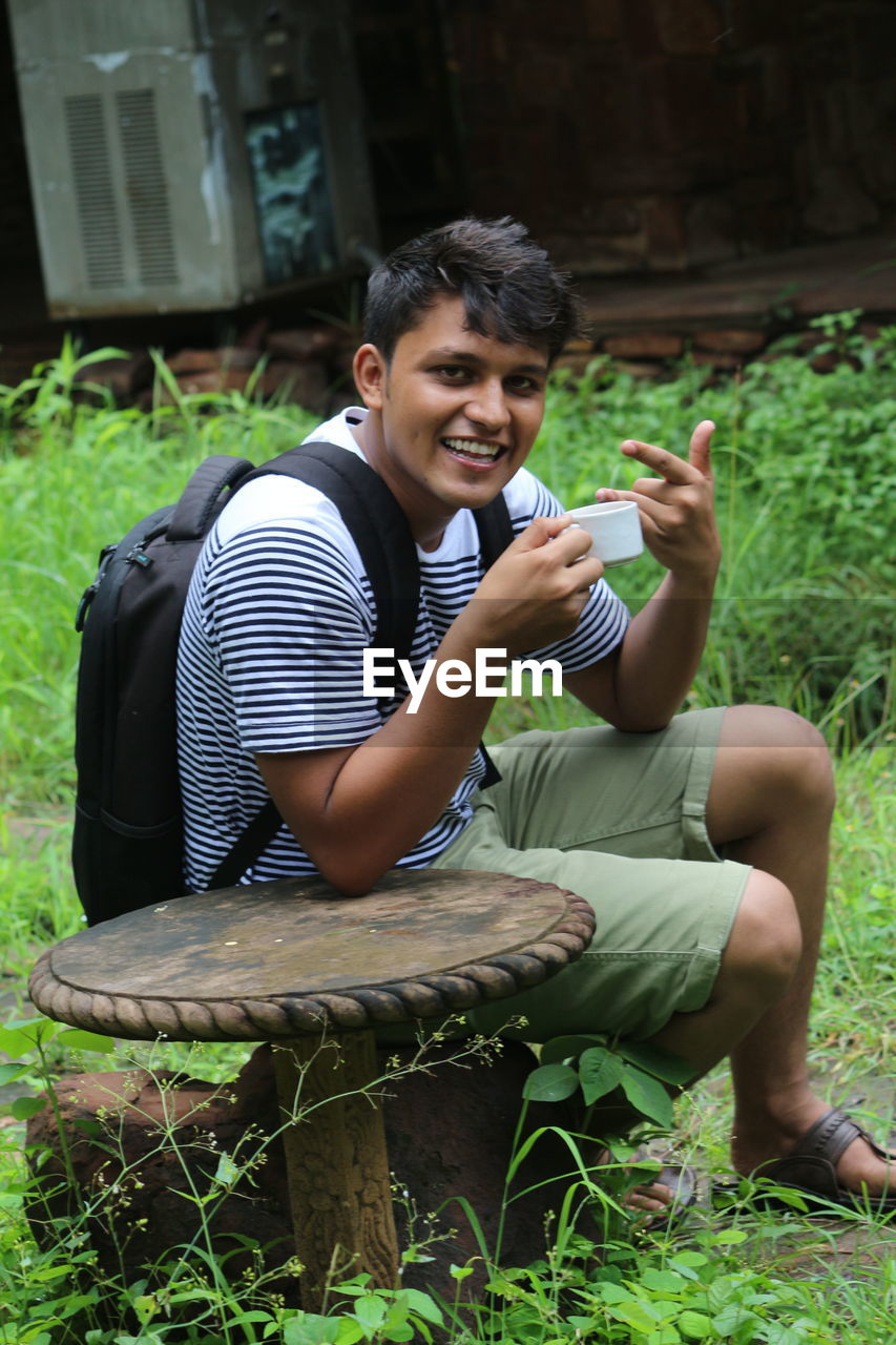 Portrait of smiling man holding tea cup while sitting on rock by table